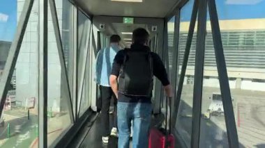 Malaga, İspanya - 24 Ocak 2025 - İki erkek yolcu güneşli bir günde valiz ve sırt çantalarını taşıyarak havaalanı jet köprüsünden bir uçağa doğru yürür. Dinamik havaalanı sahnesi.