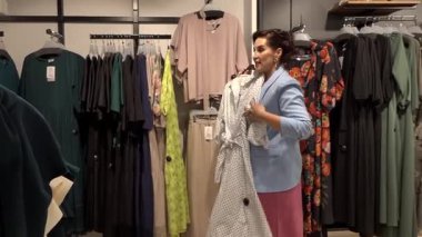 Valmiera, Letonya - 23 Nisan 2025 - Açık mavi ceketli bir kadın, asılmış giysilerle çevrili bir moda mağazasında benekli ve sarı desenli elbiseler seçiyor ve denetliyor..