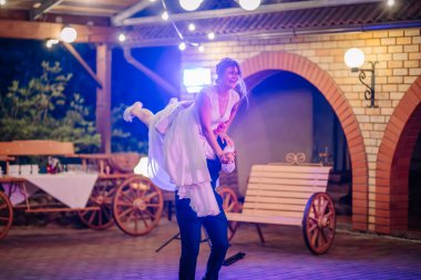 Damat düğün sırasında gelini kaldırır, beyaz bir elbise ve spor ayakkabılarla neşeyle gülerken, sıcak akşam ışıklarıyla çevrili..