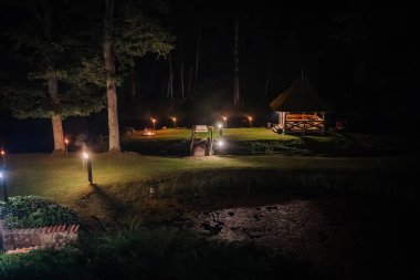 Tiki meşaleleri, şenlik ateşi, küçük bir köprü ve büyük ağaçların altında rahat ahşap bir çardak olan otlak bir bölgeyi gösteren huzurlu bir gece sahnesi..