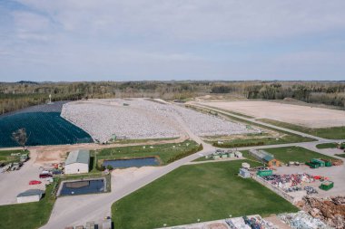 Cesis, Letonya - 28 Nisan 2025 - Arkaplanda bulutlu gökyüzünün altında atık yığınları, geri dönüşüm alanları, havuzlar, yeşil alanlar ve ormanın yer aldığı büyük bir çöp sahasının havadan görünüşü.