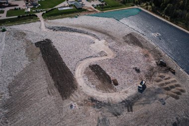 Cesis, Letonya - 28 Nisan 2025 - Dolambaçlı toprak yolu, atık yığınları, buldozerler, atık kamyonu, kapalı gölet ve yakındaki geri dönüşüm tesisi binaları olan havadan arazi manzarası.
