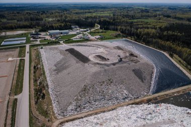 Cesis, Letonya - 28 Nisan 2025 - Orman, yollar, geri dönüşüm tesisi binaları ve parçalı bulutlu bir gökyüzü altındaki su birikintileri ile çevrili büyük bir atık sahasının havadan görünüşü.