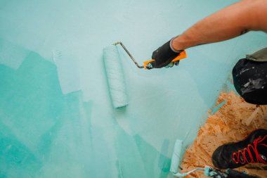Yenileme çalışmaları sırasında banyo zeminine mavi su geçirmez dolgu maddesi uygulayan kişi, yakın plan el, eldiven, silindir ve ayakkabı.