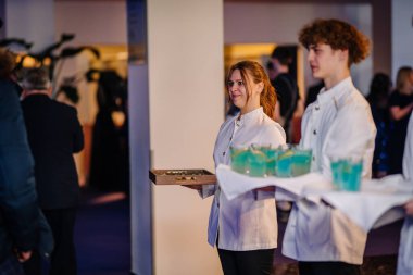Riga, Letonya - 26 Nisan 2025 - Beyaz üniformalı gülümseyen garsonlar Balta etkinliğinde mavi içki ve aperatifler ikram ederek, canlı, sıcak bir mekanda misafirlerle meşgul olurlar..