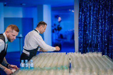 Riga, Letonya - 26 Nisan 2025 - Balta etkinlik içecek istasyonunda iki personel, serin mavi ışıklandırma altında parlayan perde arkaplanı ile konuklar için hazırlanıyor..