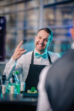 Riga, Letonya - 26 Nisan 2025 - Balta etkinliğindeki neşeli bir barmen barın arkasında parlak, parlak bir mekanda nane rengi papyon ve önlük takıyor..