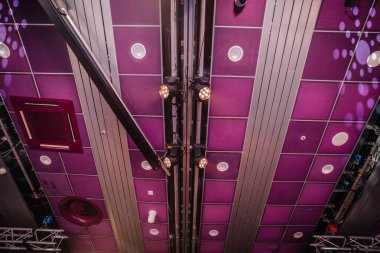 Riga, Letonya - 26 Nisan 2025 - Mor akustik panelleri, monte edilmiş spot ışıkları, havalandırma ve sahne donanımı olan, sıcak ve serin tonlarla aydınlatılmış bir etkinlik mekanının tavanı.