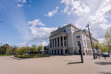 Riga, Letonya - 3 Mayıs 2025 Riga 'daki Letonya Ulusal Opera Binası dramatik bulutlar ve önünde boş bir plaza alanı bulunan mavi gökyüzü altında.