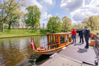 Riga, Letonya - 3 Mayıs 2025 - İnsanlar Riga Kanalı 'ndaki bir rıhtımda Letonya bayrağıyla ahşap bir gezi teknesine biniyor..