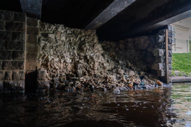 Riga, Letonya - 3 Mayıs 2025 - Suyun kenarında beton bir köprünün altında yıkılmış taş bir duvar, kentsel nehir ortamında aşınma ve çürüme belirtileri gösteriyor..