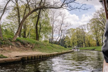 Riga, Letonya - 3 Mayıs 2025 - Bereketli yeşil park ağaçları, çimenli bankalar ve kentsel doğa ortamında berrak bir bahar gökyüzü ile çevrili küçük bir teknesi olan manzaralı bir kanal manzarası.