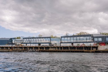 Riga, Letonya - 3 Mayıs 2025 - Nehir kıyısı boyunca inşaat halindeki modern cam bir binanın yakın görüntüsü, üzerinde iskele, ekipman ve bulutlu gökyüzü.