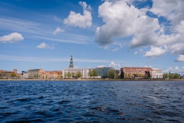 Riga, Letonya - 3 Mayıs 2025 - Daugava Nehri kıyısındaki Rigas eski kasaba manzarası, renkli tarihi binalar ve parçalı mavi gökyüzü altında kilise kuleleri