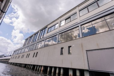 Riga, Letonya - 3 Mayıs 2025 - Geniş cam pencereli, bulutları yansıtan modern liman binası, bulutlu bir gökyüzünün altında su seviyesinden görülüyor.