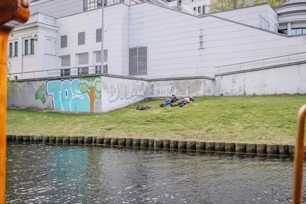 Riga, Letonya - 3 Mayıs 2025 - Duvarında grafiti olan beyaz bir binanın önündeki çimlerin üzerinde uzanan iki kişi, şehir ortamında sessiz bir anın tadını çıkarıyorlar..