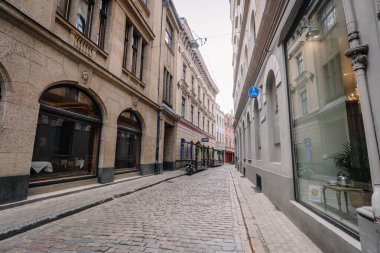 Riga, Letonya - 3 Mayıs 2025 Dar kaldırım taşı caddesi tarihi binalar, kemerli pencereler ve sakin bir Avrupa eski kasabasında yer alan küçük bir açık hava restoranı ile çevrilidir..