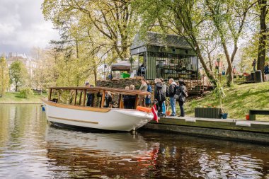 Riga, Letonya - 3 Mayıs 2025 - Bir grup insan nehir kıyısındaki küçük bir rıhtımda ahşap bir tur botuna biniyor.