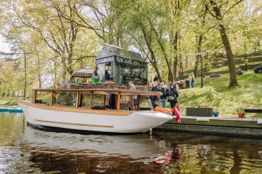 Riga, Letonya - 3 Mayıs 2025 - Bir grup insan nehir kıyısındaki küçük bir rıhtımda ahşap bir tur botuna biniyor.