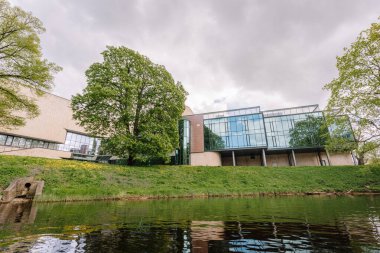 Riga, Letonya - 3 Mayıs 2025 - Nehir kenarındaki modern cam ve beton bina, yemyeşil çimenler, büyük ağaçlar ve bulutlu bir gökyüzünün altındaki sudaki yansımalarla çevrili..