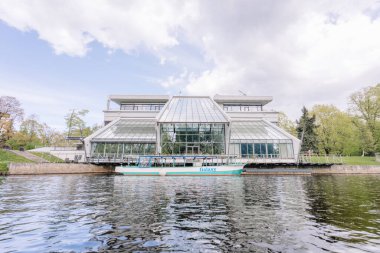 Riga, Letonya - 3 Mayıs 2025 - Su kenarındaki modern cam ve beton bina Galaxy adında bir gezi teknesi ve parktaki küçük bir deniz feneri