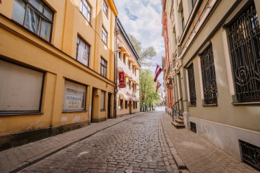 Riga, Letonya - 3 Mayıs 2025 - Eski Riga 'da tarihi binalar, Letonya bayrakları ve güneşli bir gökyüzünün altında antika bir otel tabelası bulunan dar bir kaldırım taşı caddesi.