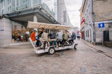 Riga, Letonya - 3 Mayıs 2025 - Old Riga 'da pedallı mobil bara binen turistler, ARKET mağazasından geçerek Letonya bayrakları ve tarihi kentsel manzaralarla döşenmiş sokaklar.