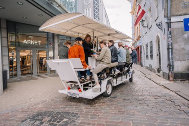 Riga, Letonya - 3 Mayıs 2025 - Bir grup insan, Litvanya bayrakları ve tarihi binaları olan bir kaldırım taşı caddesindeki ARKET mağazasından geçerek Old Riga 'da pedal gücüyle çalışan bir bara biniyor..