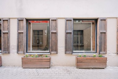Riga, Letonya - 3 Mayıs 2025 - Panjurları ve çiçek kutularıyla kaplı iki köy ahşap penceresi, eskimiş dokuyu ve kentsel cazibeyi gösteren bej bir duvar örüyor.