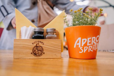 Riga, Letonya - 3 Mayıs 2025 - Aperol Spritz saksısı, baharat tutacağı albay etiketli ve arka planda bulanık kişi ile açık kafede masa kurulumu