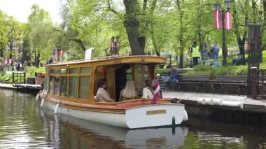 Riga, Letonya - 3 Mayıs 2025 - Lisbeth adında ahşap bir yolcu teknesi yeşil bir şehir parkı kanalına yanaşıyor ve diğer insanlar Letonya bayrakları altında kıyıda yürüyor..