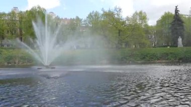 Riga, Letonya - 3 Mayıs 2025 - İnsanlar hareket halindeki bir tekneden görülen yaya köprüsü ile yeşil bir kanal kenarı parkı boyunca yürüyüp dinleniyorlar.