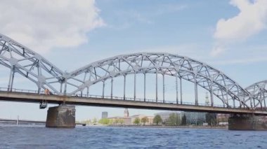 Riga, Letonya - 3 Mayıs 2025 - Riga 'daki ikonik metal demiryolu köprüsünün Daugava Nehri' ni geçerken arka planda şehir binaları ve kilise kuleleri ile detaylı görüntüsü.