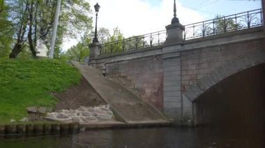 Riga, Letonya - 3 Mayıs 2025 - Taştan yaya köprüsü süslü tırabzan ve drenaj borularıyla, kanal su seviyesinden, ilkbaharda yemyeşil ağaçlarla birlikte görülür.