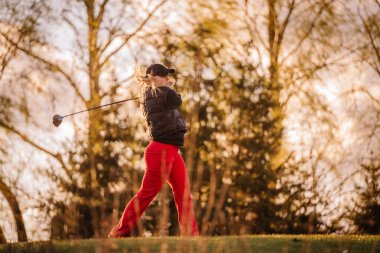 Bir kadın golfçü güçlü bir vuruşu altın saatinde tamamlar, yeşil bir sahada güvenle durur sıcak ışıkta ağaçlarla çevrili..