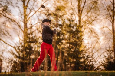 Kırmızı pantolonlu bir bayan golfçü salıncağını altın saatinde, güneşli ağaçlarla çevrili manzaralı bir sahada bitirir..