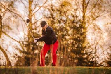 Kırmızı pantolonlu bir kadın golfçü, gün batımında yeşil bir sahada, ağaçların çerçevelediği bir sopayı sallıyor..