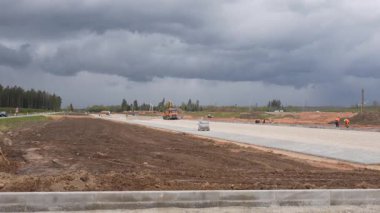 Valmiera, Letonya - 6 Mayıs 2025 - İnşaat işçileri ve makineler büyük ölçekli bir altyapı sahasında dramatik fırtına bulutları altında yeni döşenmiş bir yol bölümünde görüldü.