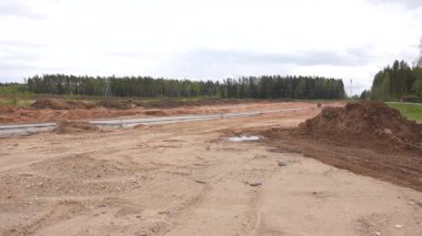 Valmiera, Letonya - 6 Mayıs 2025 - Toprak tepecikleri, erken yol temel çalışmaları ve bulutlu gökyüzü altında orman arka planına sahip kumlu bir inşaat alanı geniş görünümü.