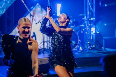 Riga, Letonya - 26 Nisan 2025 - BALTA etkinliği sırasında arkalarında canlı sahne ışıkları ve müzik aletleri ile aydınlatılan iki siyah pullu dansçı.