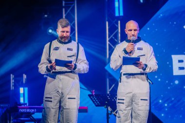 Riga, Letonya - 26 Nisan 2025 - BALTA etkinliğinde beyaz astronot kıyafetli iki adam ellerinde mikrofon ve notlar, mavi ve mor sahne ışıkları ile sahnede.