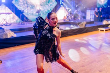 Riga, Letonya - 26 Nisan 2025 - BALTA etkinliği sırasında, canlı sahne ışıkları altında, ahşap bir dans pistinde, siyah püsküllü bir elbiseyle enerjik bir şekilde dans eder..