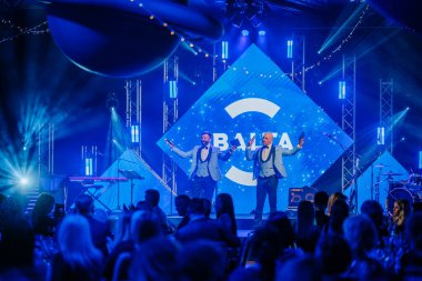 Riga, Letonya - 26 Nisan 2025 - Parlak ışıklar altında bir BALTA etkinliğinde iki parlak takım elbiseli ev sahibi, şenlikli bir atmosferde zarif bir şekilde oturan izleyicilere sesleniyor.