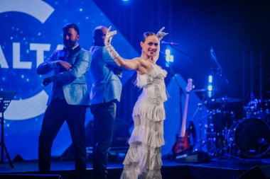 Riga, Letonya - 26 Nisan 2025 - Beyaz püsküllü elbiseli kadın dansçı, canlı bir gala gösterisi sırasında BALTA tabelası önünde iki uygun erkekle sahneye çıkıyor..