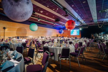 Riga, Letonya - 26 Nisan 2025 - Sallanan gezegenler, zarif yuvarlak masalar, beyaz çarşaflar, çiçekli süsler ve neşeli bir atmosferde kozmik temalı ışıklandırma ile BALTA etkinlik salonu.