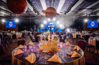 Riga, Letonya - 26 Nisan 2025 - Kozmik temalı ziyafet düzeniyle BALTA etkinliği, Satürn konsolu, zarif masalar ve büyük bir salonda canlı performansa hazır bir sahne