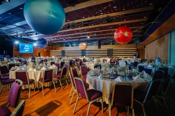 Riga, Letonya - 26 Nisan 2025 - Sallanan gezegenler, zarif yuvarlak masalar, beyaz çarşaflar, çiçekli süsler ve neşeli bir atmosferde kozmik temalı ışıklandırma ile BALTA etkinlik salonu.