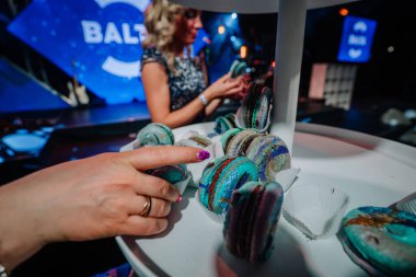 Riga, Letonya - 26 Nisan 2025 - BALTA etkinliğinde gösterilen renkli galaksi temalı badem kurabiyeleri, bir tanesini elle seçiyor ve arka planda şenlikli ışıklandırma altında bulanık bir bayan konuk seçiyor.