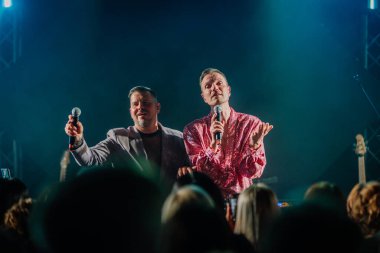 Riga, Letonya - 26 Nisan 2025 - BALTA şarkıcıları konser sırasında düet yapıyor, biri parlak pembe tişörtlü, diğeri gri takım elbiseli, karamsar sahne ışıkları altında seyircilerle takılıyor.