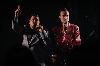 Riga, Letonya - 26 Nisan 2025 - BALTA ikilisi karanlık sahnede şarkı söylüyor, biri pembe pullu tişörtlü, biri takım elbiseli, her ikisi de dramatik, düşük ışıklı performans anında mikrofon taşıyor..